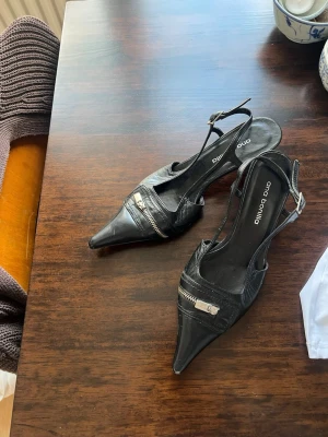 Taxklackar med silver klack - Säljer ett par svarta pumps från tidigt 2000-tal. Med silvrig klack, i skinn med spetsig tå,  öppen häl. Skorna har en justerbar rem runt ankeln och coola dragkedjedetaljer på ovansidan som ger en edgy look. Perfekta för dig som vill sticka ut lite extra. Storlek 36 men passar mig som har 37 