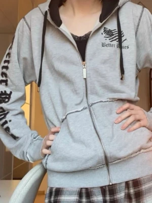 Grå zip hoodie med tryck - Grå hoodie med dragkedja från Better Bodies. Har svart tryck på bröstet och längs ena ärmen, samt svart snöre i huvan. Fickor framtill och ribbade muddar vid ärmslut och midja. Perfekt för en avslappnad streetwear-look. Ena snöret till luvan har blivit lite slitet vilket syns på sista bilden, men inget man tänker på!