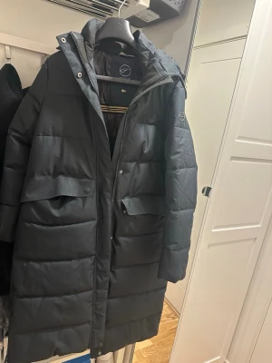 Svart lång pufferjacka från five seasons - Snygg svart lång pufferjacka med huva och stora fickor framtill. Jackan har quiltad design och dragkedja med tryckknappar. Perfekt för kalla vinterdagar och riktigt varm. Svartmelerad färg 