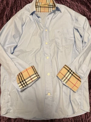 Ljusblå skjorta från Burberry  - Säljer en ljusblå skjorta från Burberry Brit med klassiskt rutigt mönster på insidan av krage och manschetter. Skjortan har bröstficka, knappar framtill och är långärmad. Perfekt för dig som gillar stilrena detaljer och ikonisk design. Har 4 jätte små hål kan finnas nått till men har inte hittat något därför säljer jag billigt
