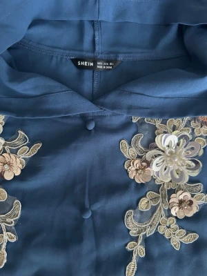 Blå klänning med spets och huva från SHEIN S - Blå klänning med huva från SHEIN, dekorerad med broderade blommor och paljetter i beige och guldtoner. Korta ärmar med spets och fransar. Knappstängning framtill och tillhörande bälte.