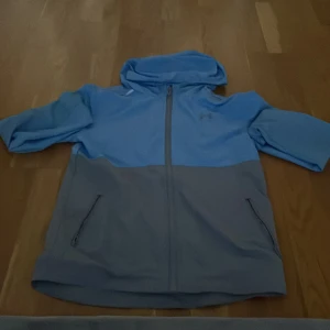 Blå och grå vindjacka från Under Armour - Sportig vindjacka från Under Armour i ljusblått och grått. Jackan har huva, dragkedja framtill och två fickor med dragkedja. Perfekt för blåsiga dagar och har en modern, avslappnad look.