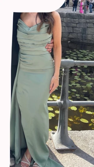 Ljusgrön långklänning med slits - Elegant ljusgrön långklänning i ett glansigt material med draperade detaljer över midjan och en hög slits på sidan. Klänningen har en figurnära passform och är ärmlös, perfekt för speciella tillfällen - balklänning. Passar 32-34