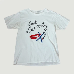 Sjukt snygg Saint Laurent tshirt  - Sjukt cool t-shirt från Saint Laurent med coolt grafiskt tryck framtill, där texten 'Saint Laurent' kombineras med röda läppar och blå detaljer. Klassisk rund hals och korta ärmar. Mjuk bomullskvalitet som känns skön mot huden. Nästan till ny, då jag inte använt den. Storlek M 