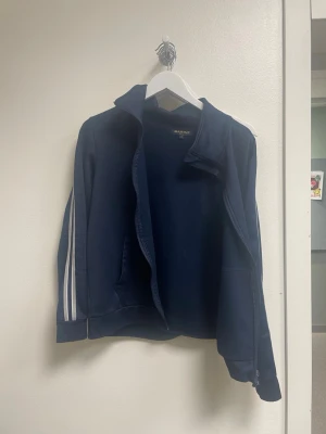 Marinblå ziptröja från Marine - Snygg marinblå ziptröja från Marine med ståkrage och vita ränder längs ärmarna. Tröjan har dragkedja framtill och två fickor. Perfekt för en sportig och avslappnad stil.