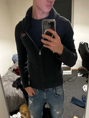 Blå stickad kofta från Hollister - Mysig blå stickad kofta från Hollister med dragkedja framtill och ribbade muddar. Koftan har hög krage och två fickor på sidorna. Perfekt för kyliga dagar och enkel att matcha med jeans eller leggings.