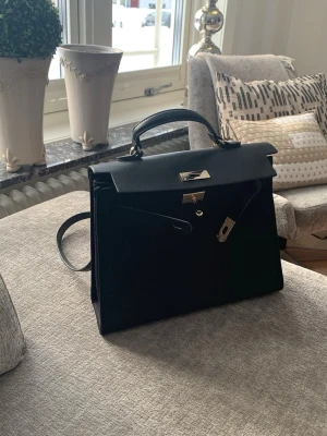 Kelly bag by Åhléns  - Stilen svart handväska från Åhléns. Väskan har avtagbar axelrem och guldfärgade metalldetaljer. Perfekt för dig som gillar tidlös design. 