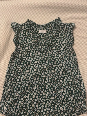 Grön blommig blus från Holly & White - Supersöt grön blus med vita småblommor från Holly & White. Blusen är ärmlös med volangdetaljer. Tunt och luftigt material. Vringad