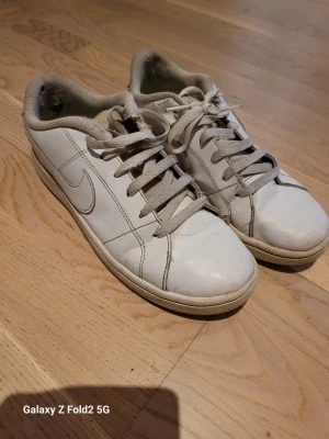 Vita Nike sneakers - Klassiska vita Nike sneakers i lågt snitt. Skorna har en enkel och stilren design med mjuk vadderad kant och ljusgrön innersula. Tillverkade i slätt lädermaterial för en clean look.