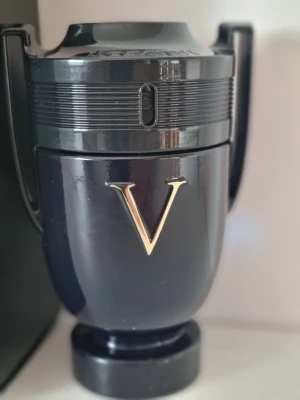 Paco Rabanne Invictus Victory parfym - Snygg parfymflaska från Paco Rabanne, modell Invictus Victory. Flaskan är svart med guldigt V på framsidan och har en unik form som en pokal med två handtag. Materialet är blank plast och glas, vilket ger en lyxig känsla.
