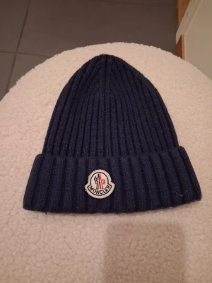 Mörkblå Moncler mössa i ullmix - Säljer en ribbstickad mörkblå Moncler mössa med uppvikt kant och klassisk logotyp framtill. Mössan är tillverkad i Italien av 70% ull och 30% kaninull, vilket gör den både mjuk och varm. Perfekt accessoar för kyliga dagar.
