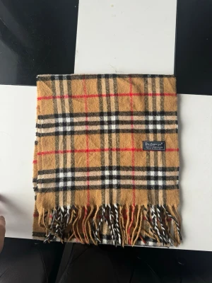 Burberrys rutig halsduk i lammull - Klassisk rutig halsduk från Burberrys i beige, svart, vitt och rött. Tillverkad i 100% lammull med fransar i ändarna och ikoniskt rutmönster. Perfekt accessoar för att lyfta din outfit med en tidlös brittisk stil.