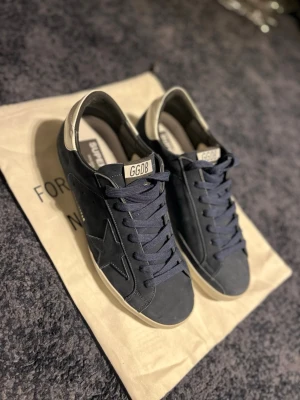 Mörkblå Golden Goose sneakers - Snygga mörkblå sneakers från Golden Goose med vit häl och GGDB-detalj på plösen. Skorna har klassisk låg modell, stjärndetalj på sidan och är gjorda i mocka och läder. Perfekta för en trendig och avslappnad stil.    Storlek 43 true to size. Använts ca 5 gånger på stan så fortfarande i grymt skick. Pris ej hugget i sten kom gärna med frågor och funderingar:)