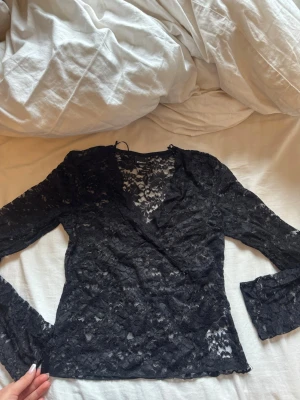 Svart spetsblus med omlott  - Snygg svart blus i genombruten spets med omlottdesign och långa ärmar. Blusen har v-ringning och är perfekt för dig som gillar en feminin och trendig stil. Materialet är lätt och stretchigt, vilket gör den bekväm att bära. Sitter som S på mig