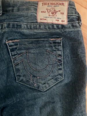 True Religion blå jeans med kontrastsöm - Säljer ett par klassiska blå jeans från True Religion med deras ikoniska kontrastsömmar på bakfickan och snygg knappdetalj fram. Jeansen har normal passform och är tillverkade i slitstarkt denimtyg. Perfekta för dig som gillar streetwear och vill sticka ut.