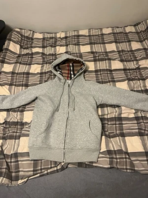 Fin Grå hoodie från Burberry med dragkedja - Säljer en grå hoodie från Burberry med dragkedja och klassiskt rutigt foder i huvan. Hoodien har två fickor framtill, justerbar dragsko i huvan och ribbade muddar. Perfekt för dig som vill ha en stilren och lyxig look med ikoniska Burberry-detaljer. Knappt använd i väldigt bra skick! Skickas samma dag eller dagen efter 