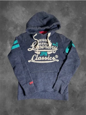 Blå Superdry hoodie med tryck - Mörkblå hoodie från Superdry med stor vit och turkos text på bröstet och två turkosa ränder på ärmarna. Klassisk känguruficka framtill, dragsko i huvan och mjukt, borstat innerfoder. Perfekt för en avslappnad streetwear-look.