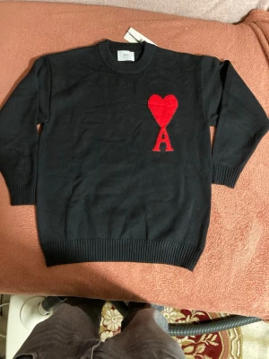 Svart stickad tröja Ami Paris hjärta - Svart stickad tröja från Ami Paris med röd hjärtdetalj och bokstaven 'A' på bröstet. Tröjan har rund halsringning, ribbade muddar och nederkant. Perfekt för dig som gillar stilrena plagg med en twist.