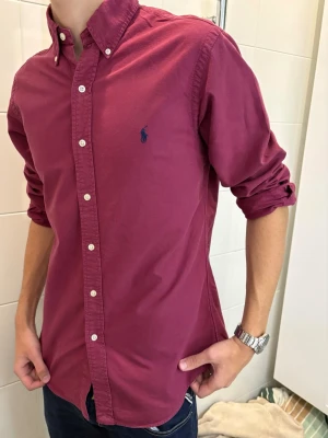 Vinröd skjorta från Ralph Lauren - Säljer en vinröd skjorta från Ralph Lauren i slim fit-modell. Skjortan har klassisk krage, långa ärmar och knappar framtill. Tillverkad i bomull och har den ikoniska loggan broderad på bröstet. Perfekt för dig som gillar stilrena plagg med snygg passform.
