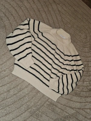 Randig stickad tröja Sisters Point - Krämvit stickad tröja från Sisters Point med svarta horisontella ränder. Tröjan har en hög hals, breda ärmar och ribbstickade muddar vid ärmslut och nederkant. Perfekt för lager-på-lager och en snygg vardagslook.