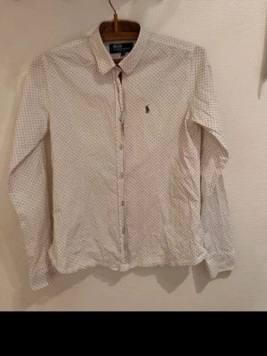Vit prickig skjorta från Ralph Lauren - Klassisk vit skjorta från Ralph Lauren med små svarta prickar och broderad logga på bröstet. Skjortan har lång ärm, knappar framtill och en tidlös krage. Perfekt för dig som gillar stilrena detaljer och ikonisk design. Är storlek L junior så är i storlek S  i vanliga 