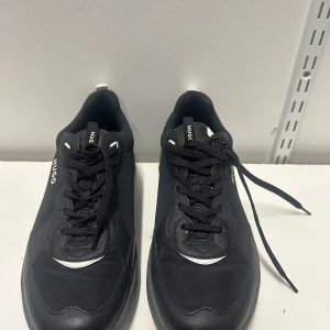 Svarta sneakers från HUGO 45 - Stilrena svarta sneakers från HUGO med vita detaljer och logga på sidan. Skorna har en chunky sula, svarta skosnören och är tillverkade i en mix av textil och syntetmaterial. Perfekta för en modern och sportig look.