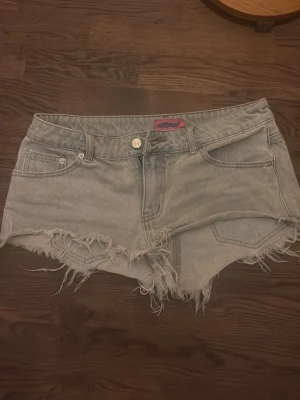 Ljusblå jeansshorts från Edikted - Säljer ett par ljusgrå jeansshorts från Edikted med råa, fransiga kanter och klassisk femficksdesign. Shortsen har låg midja och en avslappnad passform som ger en cool vibe till din outfit.