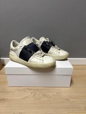 Valentino Garavani vita sneakers - Snygga vita sneakers från Valentino Garavani med bred mörk blå läderdetalj över sidan. Klassisk låg modell med rund tå och vita snören. Tillverkade i läder med diskret logga på plösen. Perfekta för dig som gillar stilrena och exklusiva sneakers. Jag fixar fler bilder vid förfrågan