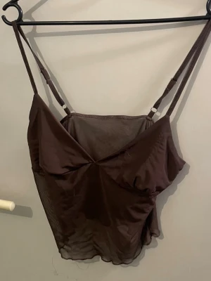 Brun mesh linnetopp med smala band - Snygg brun mesh linnetopp med smala, justerbara axelband och lätt v-ringad front. Toppen är något transparent och har en figurnära passform som ger en trendig look. Perfekt att styla med jeans eller kjol för en cool outfit.