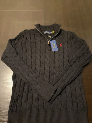 Mörkgrå kabelstickad tröja Polo Ralph Lauren - Snygg mörkgrå kabelstickad tröja från Polo Ralph Lauren med halv dragkedja och hög krage. Tröjan har det klassiska röda Polo-logot på bröstet och ribbade muddar vid ärmslut och nederkant. Perfekt för lager på lager och stilren look.