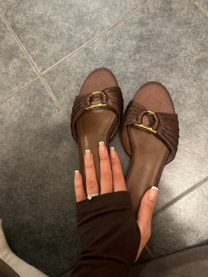 Bruna slip-in sandaler med gulddetalj - Säljer ett par bruna slip-in sandaler med bred rem i satinliknande material och guldfärgad ringdetalj framtill. Sandalerna har öppen tå och låg klack, perfekta för en trendig och avslappnad look.
