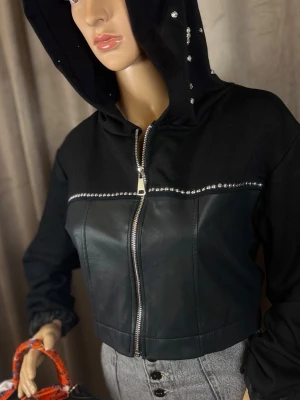 Svart cropped skinnjacka med huva - Snygg svart cropped skinnjacka med huva och dragkedja framtill. Jackan har detaljer med silverfärgade nitar längs bröstet och på huvan, vilket ger en cool och trendig look. Perfekt för dig som vill sticka ut med en edgy stil.