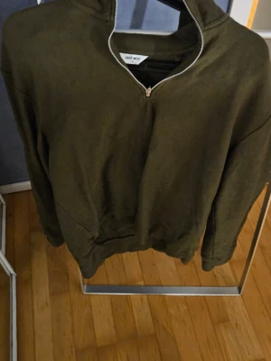 Olivgrön halvzip från East West - Snygg olivgrön halvzip från East West med halv dragkedja framtill. Tillverkad i mjukt material som känns skönt mot huden. Perfekt för dig som gillar en enkel men stilren look. Ribbade muddar vid ärmslut och nederkant.