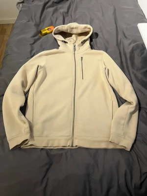 Beige hoodie från Sail Racing - Snygg beige hoodie från Sail Racing med dragkedja framtill och en bröstficka med dragkedja. Tillverkad i mjukt material och har en klassisk huva. Perfekt för en avslappnad och stilren look.
