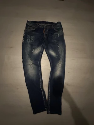 Mörkblå DSQUARED2 jeans med slitningar  - Säljer ett par mörkblå jeans från DSQUARED2 med coola slitningar och tryck på vänster lår. Jeansen har smal passform, klassiska fem fickor och en patch bak i midjan. Materialet är denim med stretch för extra komfort. Zlatan edition 