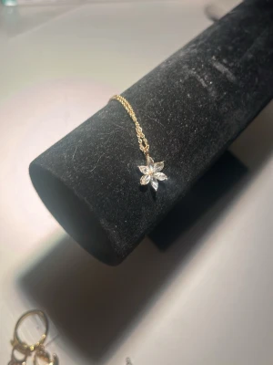 Guldarmband med blomma och kristaller - Snyggt och tunt armband i guldfärgad metall med en hängande blomma som har gnistrande, klara kristaller som kronblad. Perfekt för dig som gillar stilrena och eleganta detaljer. Lätt att matcha med andra smycken och har en enkel kedja med justerbar längd. diskutera pris 