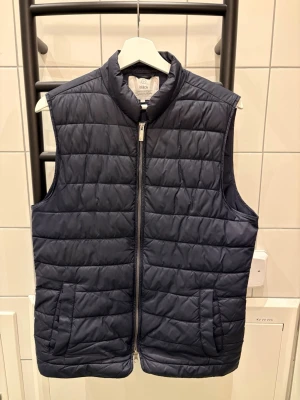 Mörkblå dunväst från Bläck - Snygg mörkblå dunväst från Bläck med quiltat mönster och hög krage. Västen har dragkedja framtill och två fickor på sidorna, knapparna har åkt av. Perfekt att slänga över en hoodie eller t-shirt för extra värme. Enkel och stilren design som funkar till det mesta.