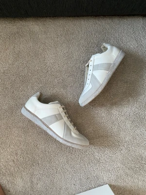 Maison Margiela White Paint Gats - Stilrena Maison Margiela sneakers i vitt skinn med grå mockadetaljer och klassisk gummisula. Modellen har snörning och en låg profil som passar till många olika stilar. Perfekta för dig som gillar minimalistisk design och neutrala färger. Innersula 28,5cm