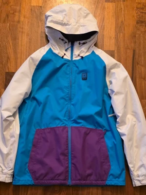 Rip Curl snowboardjacka - size Xlarge  - Snowboardjacka från Rip Curl med blå front, vita ärmar och huva samt lila fickor. Jackan har dragkedja, huva och stora fickor framtill. Perfekt för dig som gillar streetstyle och vill sticka ut med en retrolook.