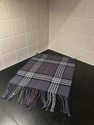 Mörkgrå halsduk med rutigt mönster - Stilren halsduk i mörkgrått med klassiskt rutigt mönster i vitt, svart och röda linjer. Halsduken har fransar i ändarna och passar perfekt för att ge din outfit en snygg touch. Materialet känns mjukt och behagligt mot huden. Liknade burberry 