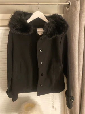 Kappa Ginatricot - Svart kappa med stor fluffig huva kantad av svart fuskpäls. Jätte bra skick  Den är i storlek M men passar även S. Använd fåtal gånger. Jackan har knappar framtill, två fickor och snygga detaljer vid ärmsluten. köpt för 500kr på Gina.