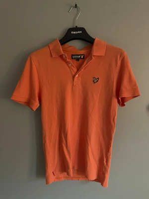 Orange pikétröja från Lyle & Scott - Snygg orange pikétröja från Lyle & Scott med klassisk krage och korta ärmar. Tröjan har knappar framtill och den ikoniska gula fågeln broderad på bröstet. Perfekt för dig som gillar stilrena och färgglada plagg.