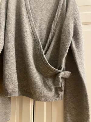 Grå omlottkofta från Kappahl - Mysig grå omlottkofta från Kappahl med knyt i sidan och djup v-ringning. Koftan är stickad och har långa ärmar, perfekt för lager-på-lager-stil under kyliga dagar. Använd fåtal gånger💗💗