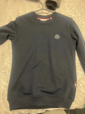 Mörkblå sweatshirt från Moncler - Snygg mörkblå sweatshirt från Moncler med diskret logotyp på bröstet och ribbade muddar vid ärmslut och nederkant. Tröjan har en klassisk rund hals och är tillverkad i mjuk bomull, perfekt för en chill och stilren look.