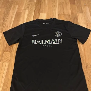 Svart PSG x Balmain fotbollströja Nike S - Svart fotbollströja från Nike i samarbete med PSG och Balmain Paris. Tröjan har korta ärmar, rund hals och är tillverkad i lätt, ventilerande material (Nike Aeroswift). Framsidan har PSG-logga och Balmain Paris-tryck i regnbågsskimrande färg.