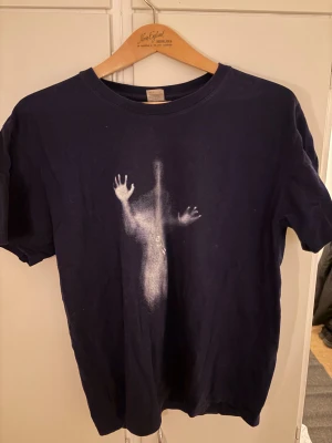 Mörkblå t-shirt med tryck - Säljer en mörkblå t-shirt med ett unikt vitt tryck på framsidan som föreställer en siluett med händer. T-shirten har rund hals och korta ärmar, perfekt för dig som gillar coola och lite mystiska motiv. Materialet känns mjukt och bekvämt mot huden.