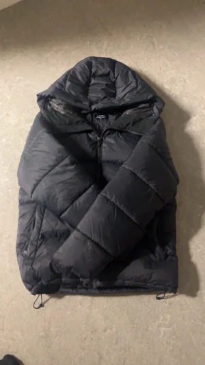 Svart pufferjacka från Everest - Säljer en svart pufferjacka från Everest med huva och dragkedja. Jackan är quiltad och har en klassisk oversized look, perfekt för kalla dagar. Märket syns på ärmen och jackan har justerbara snören nedtill för extra komfort.