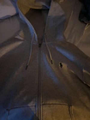 Grå hoodie från Polo Ralph Lauren - Snygg grå hoodie från Polo Ralph Lauren med dragkedja och huva med snörning. Klassisk logga broderad på bröstet. Mjuk bomullskvalitet och praktiska fickor framtill. Perfekt för chill dagar och enkel att matcha med jeans. Den är som ny. 