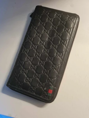 Svart plånbok från Gucci i skinn - Snygg svart plånbok från Gucci i äkta skinn med präglat GG-mönster över hela utsidan. Plånboken har dragkedja runtom, flera kortfack och ett separat fack med dragkedja inuti. Klassisk Gucci-detalj med rödgrön rand i hörnet.