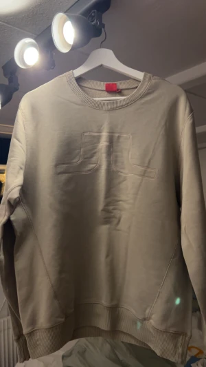 Beige sweatshirt från J.Lindeberg - Snygg beige sweatshirt från J.Lindeberg i storlek L. Tröjan har ett J.Lindeberg märke över bröstet, rund hals och ribbade muddar vid ärmslut och nederkant. Tillverkad i mjuk bomull för skön komfort. Perfekt för chill dagar och enkel att matcha.